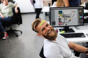 Smiling man with satisfying jobs sitting at a desk at work with post-it that says Be Happy