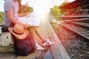 Person sitting on a bench at a train station with a camera, hat and backpack looking for somewhere to travel on holiday